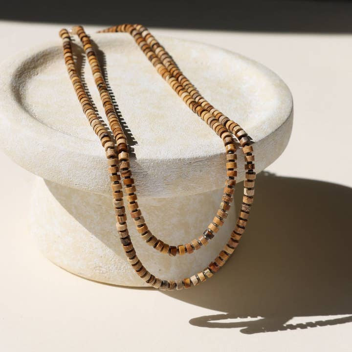 The Dune Beaded Necklace by Token Jewelry, featuring jasper gemstones, is elegantly draped over a round, textured off-white pedestal in sunlight.