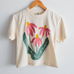 An ivory Comfort Colors boxy crop tee from Ramble & Company, named “Sow the Seeds,” featuring a graphic of three pink flowers with green leaves, hangs on a wooden hanger against a white wall.