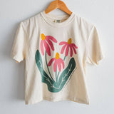 An ivory Comfort Colors boxy crop tee from Ramble & Company, named “Sow the Seeds,” featuring a graphic of three pink flowers with green leaves, hangs on a wooden hanger against a white wall.