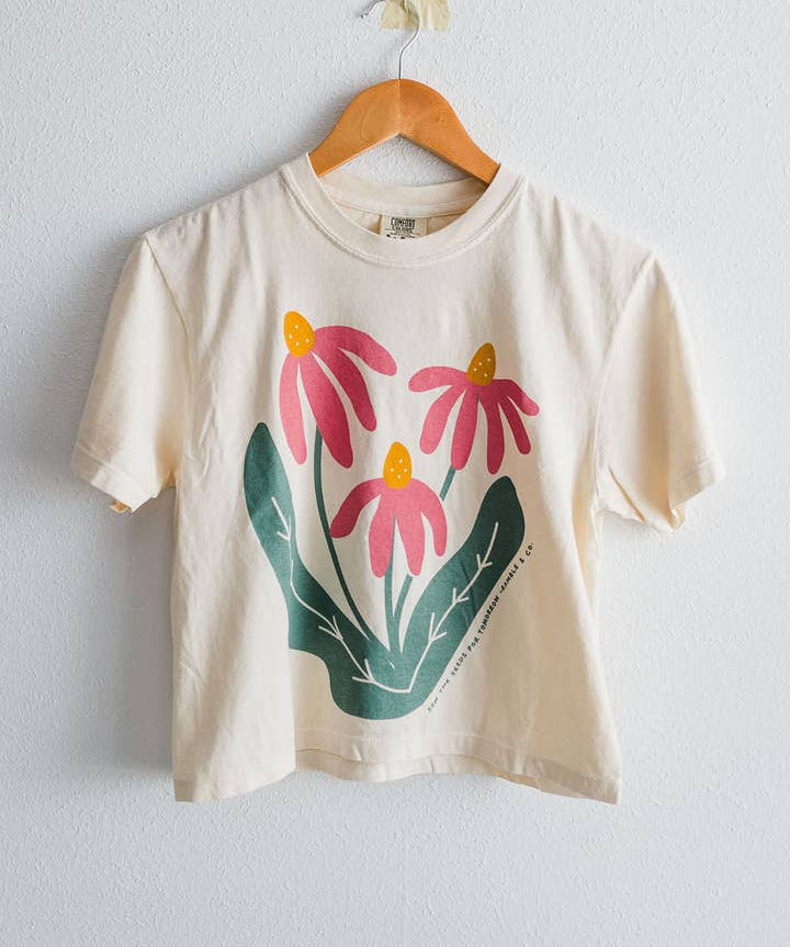 An ivory Comfort Colors boxy crop tee from Ramble & Company, named “Sow the Seeds,” featuring a graphic of three pink flowers with green leaves, hangs on a wooden hanger against a white wall.