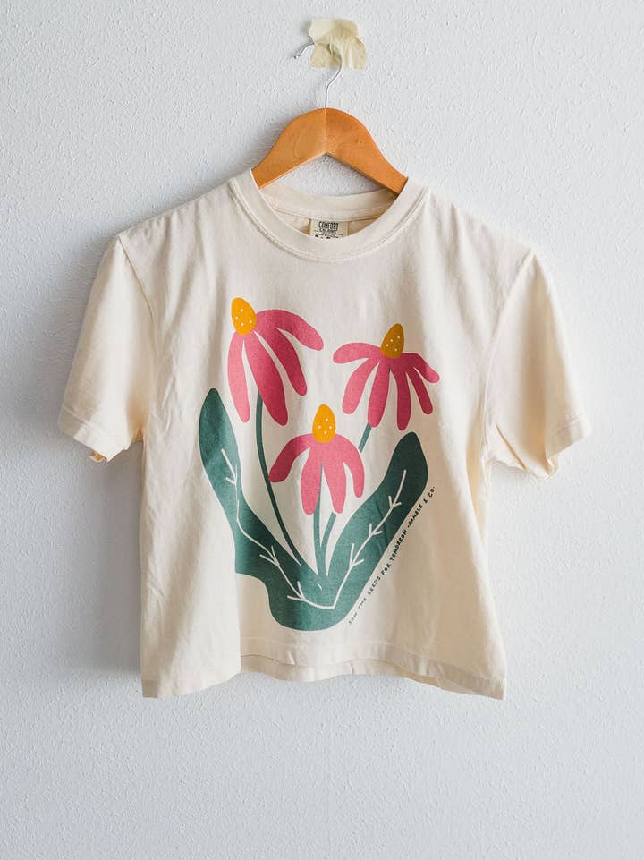An ivory Comfort Colors boxy crop tee from Ramble & Company, named “Sow the Seeds,” featuring a graphic of three pink flowers with green leaves, hangs on a wooden hanger against a white wall.