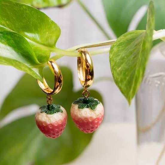 The Berry Juicy Hoops by Peter and June feature 18k gold-plated hoops with strawberry-shaped pendants, hanging from a green plant stem as their ethically sourced, water-resistant design glimmers against blurred leaves and glass in the background.