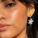 Close-up of a person's face wearing Peter and June's Earth Angel Orchid earrings—nickel free, 18k gold plated hoops featuring a blue and white hand-painted ceramic flower pendant.