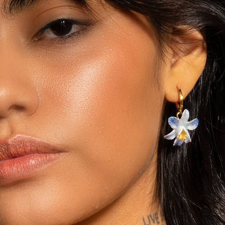 Close-up of a person wearing the Earth Angel Orchid Earrings by Peter and June—18k gold plated hoops with a hand-painted blue and white ceramic flower pendant. Only the lower half of the face and ear are visible.