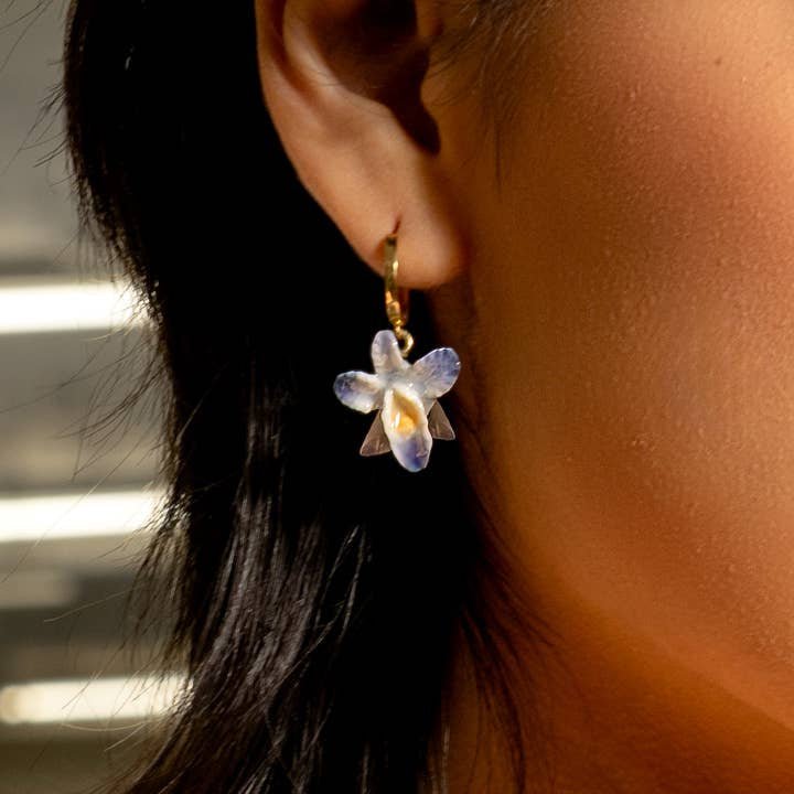 Close-up of a person wearing Peter and June's Earth Angel Orchid earrings featuring an 18k gold plated hoop and a purple and white hand-painted ceramic flower pendant.