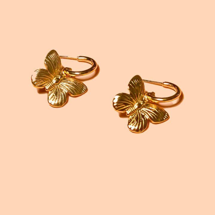 The Peter and June "I'll Fly Away Hoops" are nickel-free 18k gold plated hoop earrings with butterfly pendants, displayed on a light peach background.