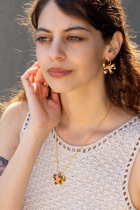 A woman wearing a crocheted beige top, Peter and June Knotty Hoops earrings, and a matching ethically sourced gold flower pendant necklace.