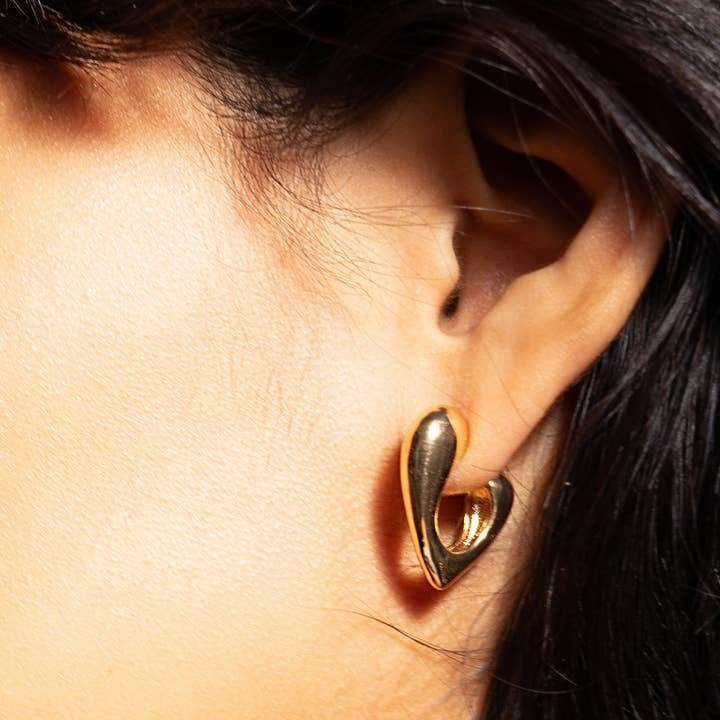 Close-up of a person's ear wearing Peter and June's Phoebe Hoops—shiny, abstract-shaped, 18k gold plated earrings that are nickel free and water resistant—against a neutral background.