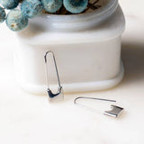 Peter and June's Sid Safety Pin Earrings, rhodium silver plated, are shown on a white surface—one leaning upright against a white container with dried blue flowers in the background.
