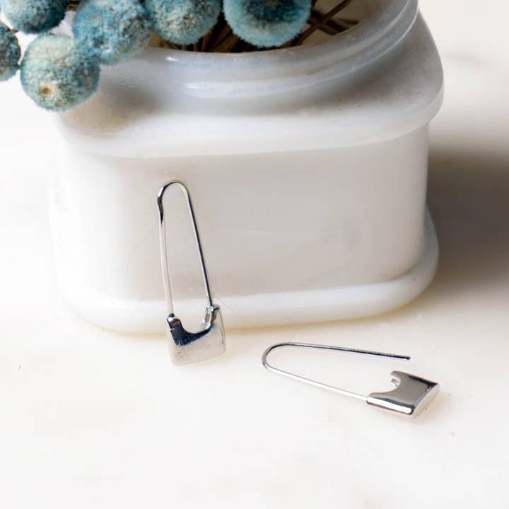 Peter and June's Sid Safety Pin Earrings, rhodium silver plated, are shown on a white surface—one leaning upright against a white container with dried blue flowers in the background.