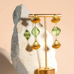 The "Under the Boardwalk" earrings by Peter and June feature gold and green geometric drops with handmade lampwork glass, shown on a gold stand before a stone and beige backdrop.