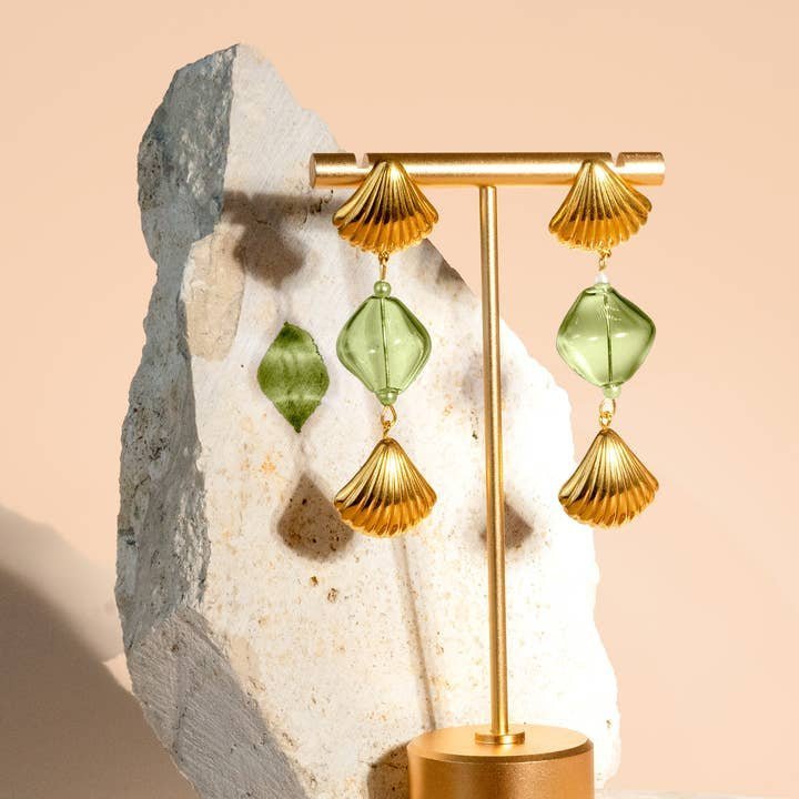 The "Under the Boardwalk" earrings by Peter and June feature gold and green geometric drops with handmade lampwork glass, shown on a gold stand before a stone and beige backdrop.