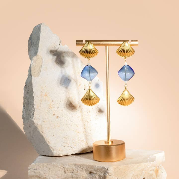 The "Under the Boardwalk" earrings by Peter and June feature gold and blue lampwork glass with 18k gold-plated accents, displayed on a gold stand against light stone props and a beige background.