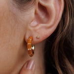 A close-up of a woman's ear with a small gold stud in the upper lobe and a larger orange enamel and gold Vintage Hoop earring from JaxKelly in the lower lobe.
