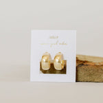 A pair of JaxKelly Earrings | Wide Hoop displayed on a white card with gold text, standing upright on a beige surface next to a brown stone.