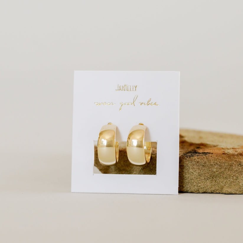 JaxKelly Wide Hoop Earrings displayed on a white card with gold "JaxKelly" and "wear good vibes" text, set before a stone block. These chunky gold statement earrings are perfect for adding a bold touch to your style.