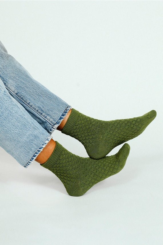 Person wearing Tailored Union Eben Ankle Sock, green midweight socks with a knitted sweater design, paired with light blue jeans, sitting with legs slightly crossed against a plain light background.