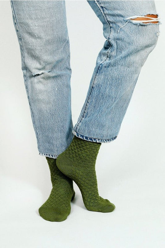 Person wears light blue ripped jeans and Eben Ankle Sock by Tailored Union, a textured green midweight ankle sock, standing with legs crossed against a plain background.