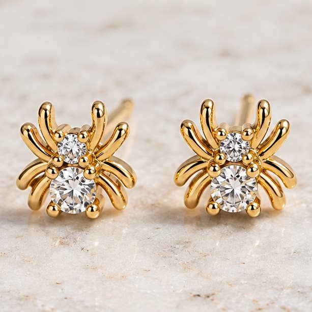 Mesa Blue Tiny Cz Spider Stud Earrings feature multiple legs and two clear round cubic zirconia gemstones on each earring, crafted from tarnish-resistant 18K gold plated metal. Displayed on a light stone surface, these unique studs from Mesa Blue offer an elegant touch and lasting shine.