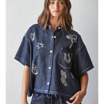 A woman is wearing the By Together Denim Embroidered short-sleeved, buttondown collar top with white western motifs and matching dark denim pants, standing against a plain light background.