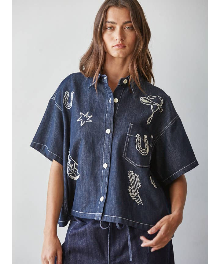 A woman is wearing the By Together Denim Embroidered short-sleeved, buttondown collar top with white western motifs and matching dark denim pants, standing against a plain light background.
