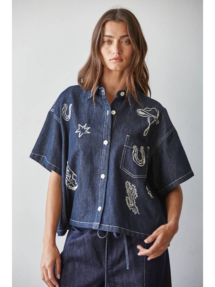A woman is wearing the By Together Denim Embroidered short-sleeved, buttondown collar top with white western motifs and matching dark denim pants, standing against a plain light background.