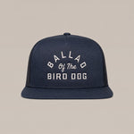The Arch Logo Hat by Ballad Originals is a navy blue trucker cap featuring a stylish arch logo and white embroidered “BALLAD OF THE BIRD DOG” text on the front, showcased against a plain beige background.