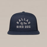 The Arch Logo Hat by Ballad Originals is a navy blue trucker cap featuring a stylish arch logo and white embroidered “BALLAD OF THE BIRD DOG” text on the front, showcased against a plain beige background.