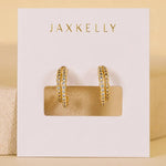 The Double Champagne Hoop earrings by JaxKelly feature gold double hoops adorned with clear pave stones, elegantly displayed on a white “JaxKelly” card against a neutral background; these hypoallergenic earrings blend everyday comfort with sophisticated style.