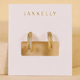 The Double Champagne Hoop earrings by JaxKelly feature gold double hoops adorned with clear pave stones, elegantly displayed on a white “JaxKelly” card against a neutral background; these hypoallergenic earrings blend everyday comfort with sophisticated style.