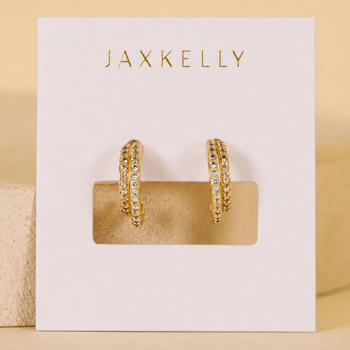 The Double Champagne Hoop earrings by JaxKelly feature gold double hoops adorned with clear pave stones, elegantly displayed on a white “JaxKelly” card against a neutral background; these hypoallergenic earrings blend everyday comfort with sophisticated style.