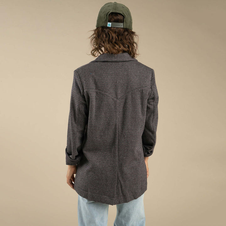 A person stands with their back to the camera wearing the Eleanor Relaxed Western Blazer by Sendero Provisions Co., styled with light blue jeans and a green baseball cap against a plain beige background.
