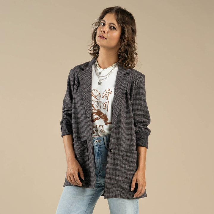 A woman wears the Eleanor Relaxed Western Blazer by Sendero Provisions Co. over a graphic tee and light blue jeans, standing against a plain beige background.