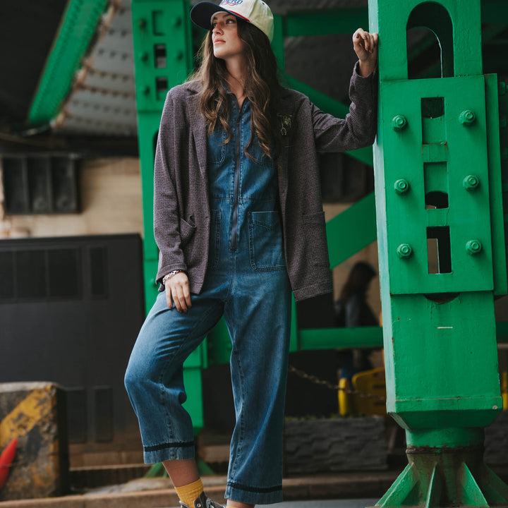 Wearing the Eleanor Relaxed Western Blazer by Sendero Provisions Co., a person in a denim jumpsuit and baseball cap leans against a green metal structure in an urban setting.