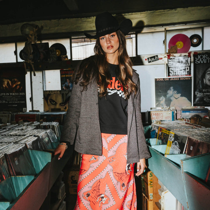 Wearing the Eleanor Relaxed Western Blazer by Sendero Provisions Co., a woman in a cowboy hat, black graphic tee, and red patterned skirt browses between rows of vinyl records in a music store.