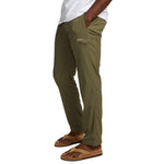 A person stands with hands in pockets, wearing olive green Howler Brothers Electrolight Tech Pants with a side zipper pocket, a white shirt, and tan sandals.