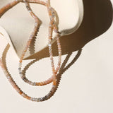 The Ember Moonstone Necklace by Token Jewelry, with delicate 14k gold fill accents and handmade multicolored beads, is draped over a curved, light-colored ceramic surface against a cream background.