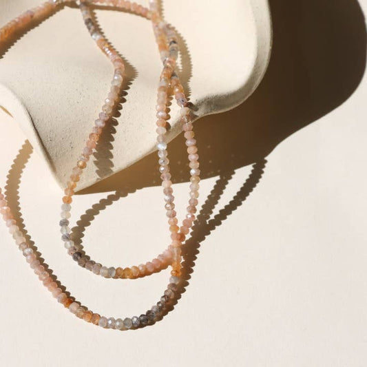 The Ember Moonstone Necklace by Token Jewelry, with delicate 14k gold fill accents and handmade multicolored beads, is draped over a curved, light-colored ceramic surface against a cream background.