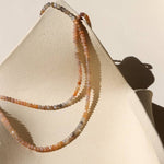 The Ember Moonstone Necklace by Token Jewelry, featuring small faceted moonstones in neutral tones, is displayed on a textured off-white ceramic dish in natural light, highlighting its unique handmade charm.