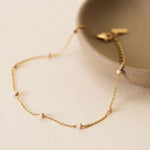 The JaxKelly Anklet in Petal Pink features a delicate diamond-cut gold chain with small pink beads, shown draped over a beige ceramic dish. Hypoallergenic for sensitive skin.