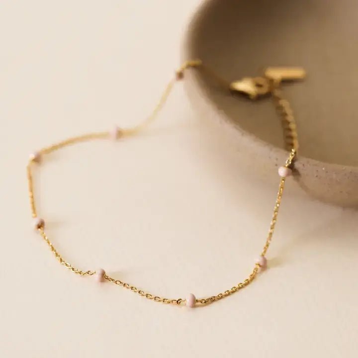 The JaxKelly Anklet in Petal Pink features a delicate diamond-cut gold chain with small pink beads, shown draped over a beige ceramic dish. Hypoallergenic for sensitive skin.