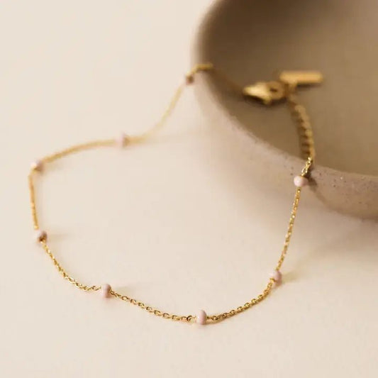 The JaxKelly Anklet | Petal Pink features a delicate diamond cut chain with small pink beads, shown beside a beige ceramic dish on a light surface—tarnish-free for lasting shine.