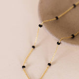 The Enamel Gold Necklace - 18k Gold Plated - Black by JaxKelly is draped over the edge of a light-colored ceramic bowl, showing off its delicate, tarnish-free design and small black beads.