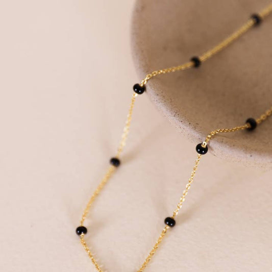 The Enamel Gold Necklace - 18k Gold Plated - Black by JaxKelly features delicate black beads on a fine chain, elegantly draped over a light ceramic dish for a refined accent to any outfit.