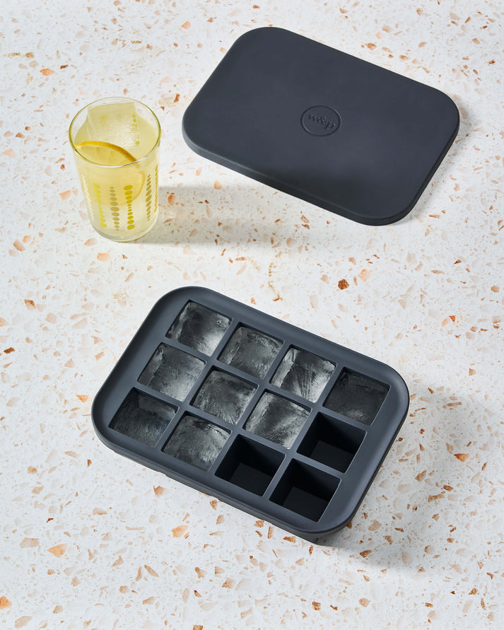 The W&P Everyday Ice Tray, a premium silicone ice cube tray with a lid, sits on a speckled surface next to a glass of yellow drink with ice.