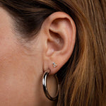 Close-up of a person’s ear adorned with two earrings from JaxKelly: a small, square-shaped stud on the earlobe and the JaxKelly Everyday Silver Hoop Earring hanging below. Both pieces are hypoallergenic and designed to be tarnish free for lasting shine.