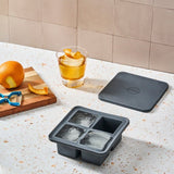 The W&P Extra Large Ice Cube Tray, made of BPA-free silicone, sits on a speckled countertop with three ice cubes, a drink garnished with orange peel, an orange, a peeler, and the tray’s lid.