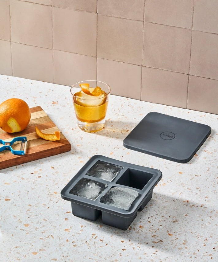 The W&P Extra Large Ice Cube Tray, made of BPA-free silicone, sits on a speckled countertop with three ice cubes, a drink garnished with orange peel, an orange, a peeler, and the tray’s lid.