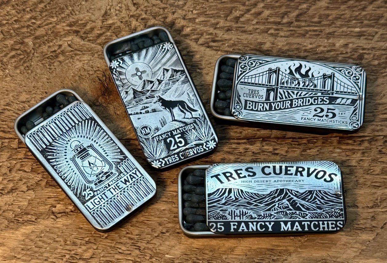 Four Fancy Matches | Tres Cuervos matchboxes by Tres Cuervos, each adorned with intricate black and white designs and bold text such as “BURN YOUR BRIDGES” and “LIGHT THE WAY,” are displayed on a wooden surface, capturing the adventurous spirit of the American West.