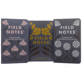 The Field Notes Fall Flowers 3-Pack includes three 48-page memo books with early-20th-century-inspired botanical covers, featuring pink, yellow, and white fall flowers on dark backgrounds, standing upright side by side.